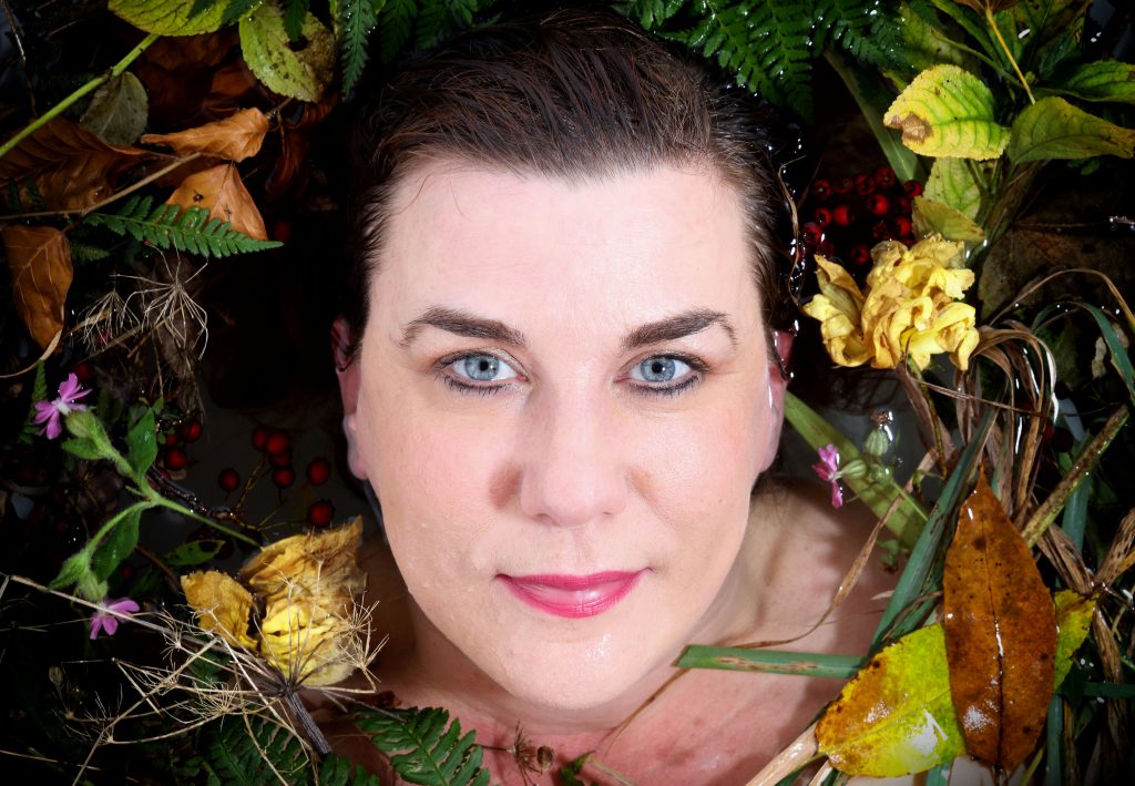 Headshot of poet Kate Fox, she is surrounded by foliage.
