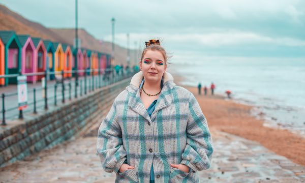 Lizzie Lovejoy stands on Saltburn beach (with the brightly coloured beach huts in the background). They are wearing a checked coat, bright eyeshadow, and their hair is tied up in a bun.