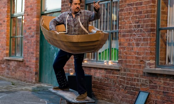 A man stands on a board balanced on a small barrel shaped object. He is wearing a cardboard boat hanging by straps over his shoulders.