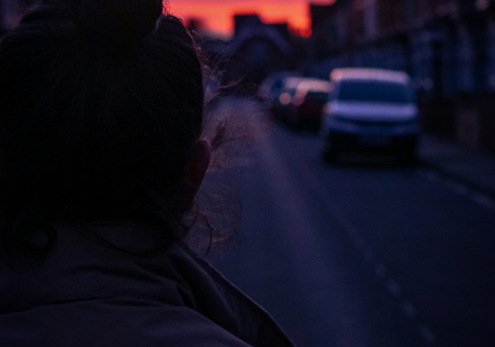 A figure in shadow in the foreground looks down a street lined with terraced houses and parked cars, towards a pink and orange sky.