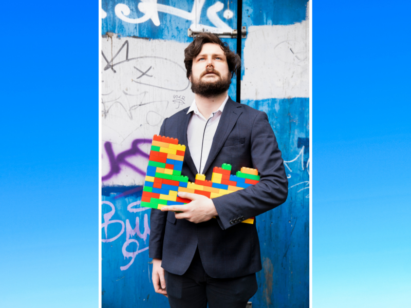 A young man with thick dark hair and a beard, wearing a suit, stands against a graffitied wall, looking upwards. He is holding a 'wall' of multi-coloured Lego pieces.
