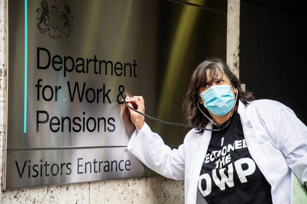 A person dressed in a doctor's coat and wearing a face mask holds a stethoscope to a metal sign on a wall reading 'Department for Work & Pensions / Visitors Entrance'