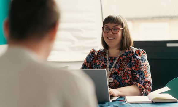 A woman in her mid thirties, with shoulder length hair and red framed glasses, sits at a table working on a laptop. She is smiling at other colleagues around the table.