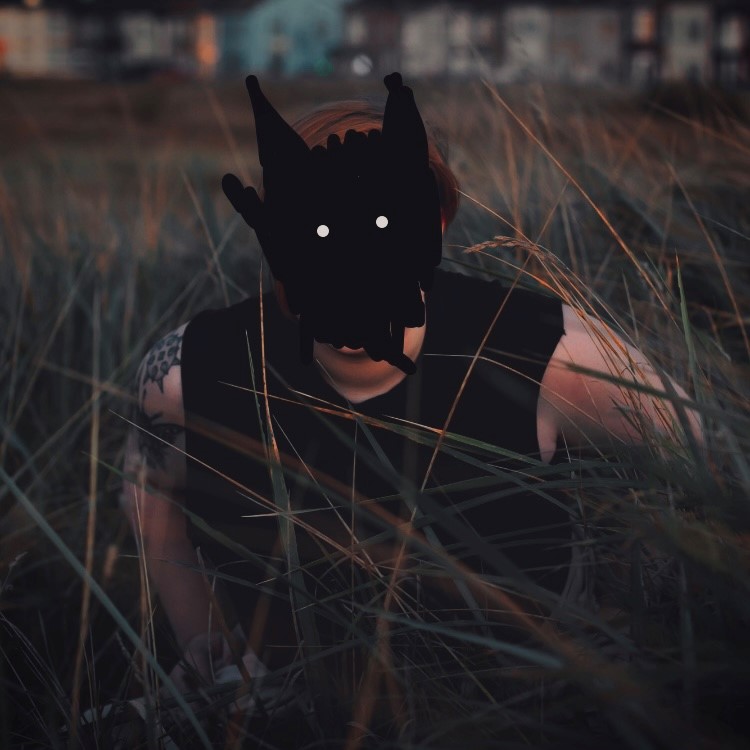 Performer Audrey Cook crouched in long grass with their face covered by a black mask that has been drawn over the photo