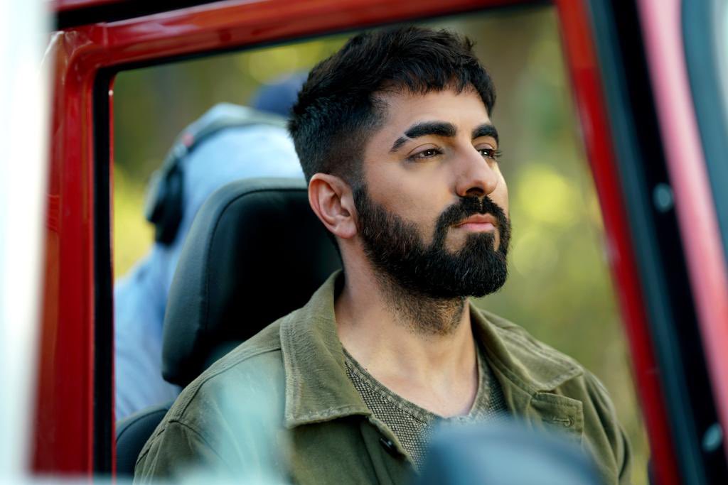 A south Asian man with dark hair and a beard sits in the front seat of a car, looking thoughtfully into the distance.
