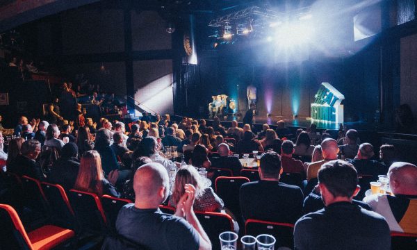 A crowded room lit by bright white stage lights, the audience are all laughing and clapping, on the stage is a 3d prop which reads Beat the Gong.