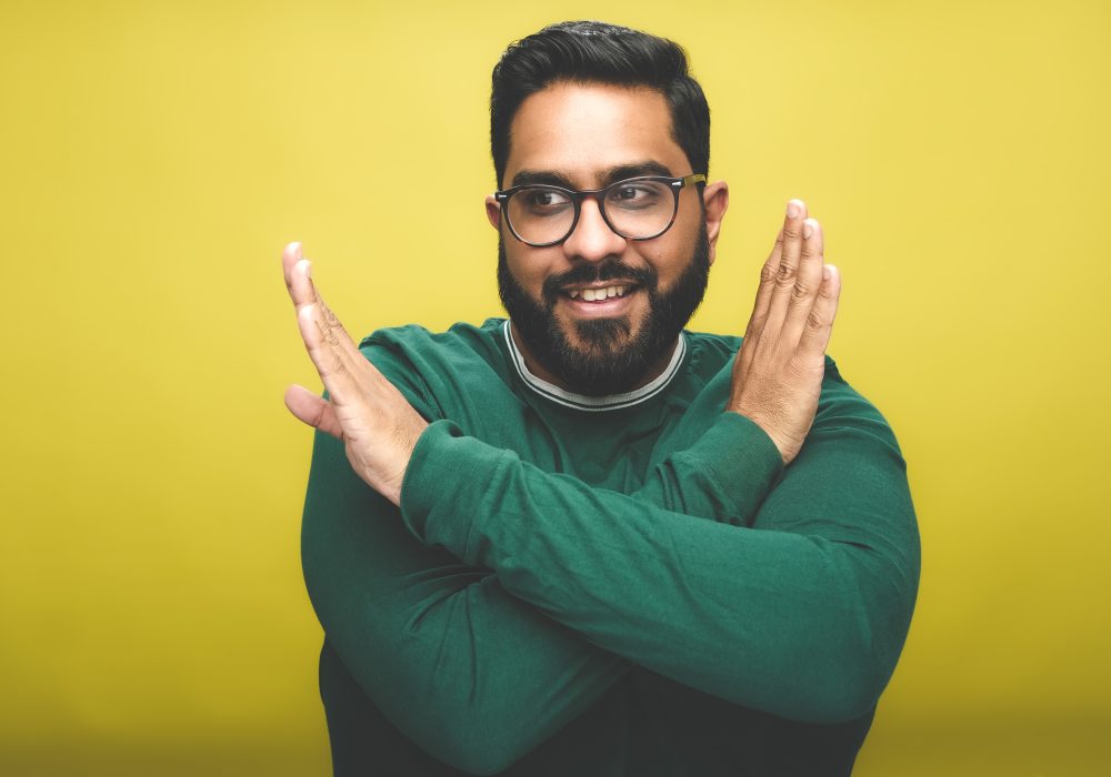 Comedian Eshaan Akbar stands in front of a yellow backdrop. He wears a green jumper, and has both of his arms across his body to make an X shape, he is looking off to the side of the camera and is smiling.