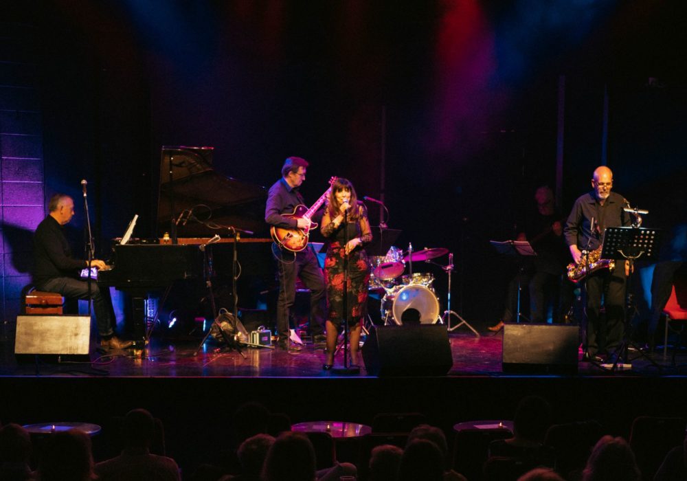A jazz band on a lit stage, their female singer is centre stage looking out at a crowded room. The audience is visible in the bottom of the image.