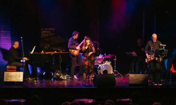 A jazz band on a lit stage, their female singer is centre stage looking out at a crowded room. The audience is visible in the bottom of the image.