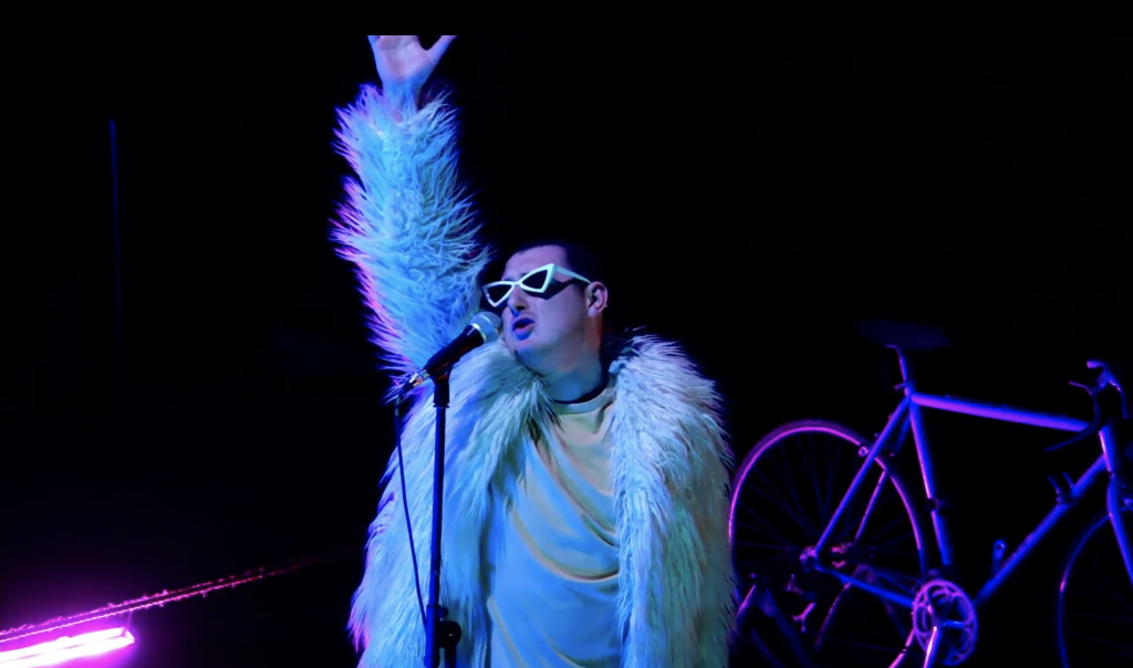A person wearing sunglasses on a dark stage. A bicycle is behind them.