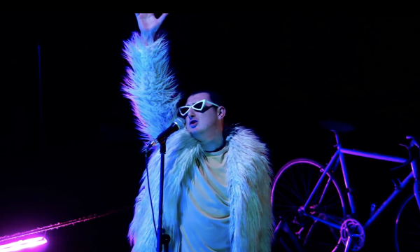 A person wearing sunglasses on a dark stage. A bicycle is behind them.