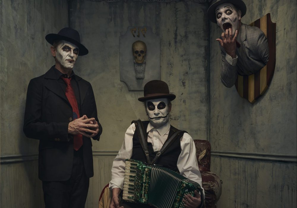 Two men are present in a dingy grey room, the man on the left is standing with his hands tented, and the other man is sitting in a cracked armchair holding a green accordian. Behind them both is a skull mounted on the wall. To the right of the photo a third man, with only his head and shoulders visible appears to be mounted on the wall. His mouth and eyes are wide open as if in shock. All three men have black and white face paint on, and they are all smartly dressed in suits and ties.