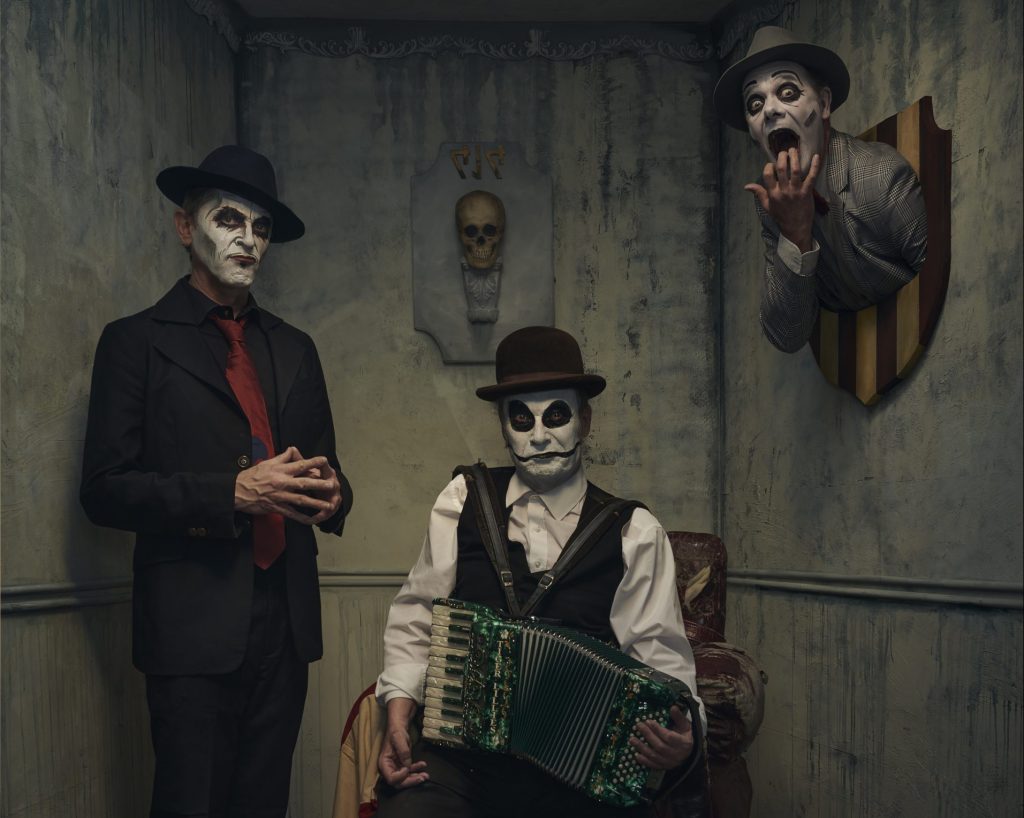 Two men are present in a dingy grey room, the man on the left is standing with his hands tented, and the other man is sitting in a cracked armchair holding a green accordian. Behind them both is a skull mounted on the wall. To the right of the photo a third man, with only his head and shoulders visible appears to be mounted on the wall. His mouth and eyes are wide open as if in shock. All three men have black and white face paint on, and they are all smartly dressed in suits and ties.