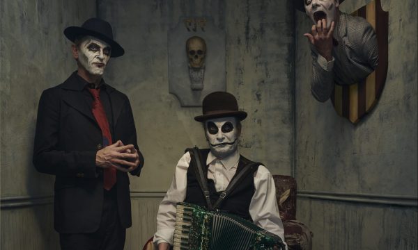 Two men are present in a dingy grey room, the man on the left is standing with his hands tented, and the other man is sitting in a cracked armchair holding a green accordian. Behind them both is a skull mounted on the wall. To the right of the photo a third man, with only his head and shoulders visible appears to be mounted on the wall. His mouth and eyes are wide open as if in shock. All three men have black and white face paint on, and they are all smartly dressed in suits and ties.