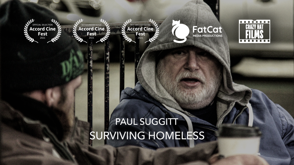Text reading 'Paul Suggitt Surviving Homeless', Crazy Hat Films, and Accord Cine Fest selections. A grey-bearded man in a grey hoodie and blue jacket is speaking to a man in a brown jacket and black hat.