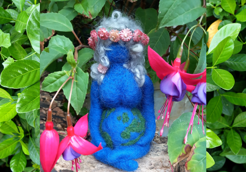 A photo of a blue needle felted mother earth sculpture, she is cradling a bump that represents the earth, her hair is grey and curled and she wears a tiny pink flower crown. The sculpture is sitting cross-legged in front of a green bush, and there are two fuschia plants, one on either side of the sculpture.