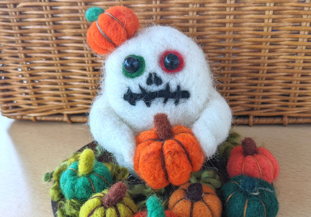 A needle felt sculpture of a ghost, it is sitting in a pumpkin patch, with one orange pumpkin on top of it's head, and it is holding another orange pumpkin.