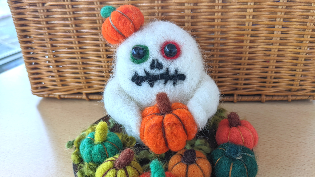 A needle felt sculpture of a ghost, it is sitting in a pumpkin patch, with one orange pumpkin on top of it's head, and it is holding another orange pumpkin.