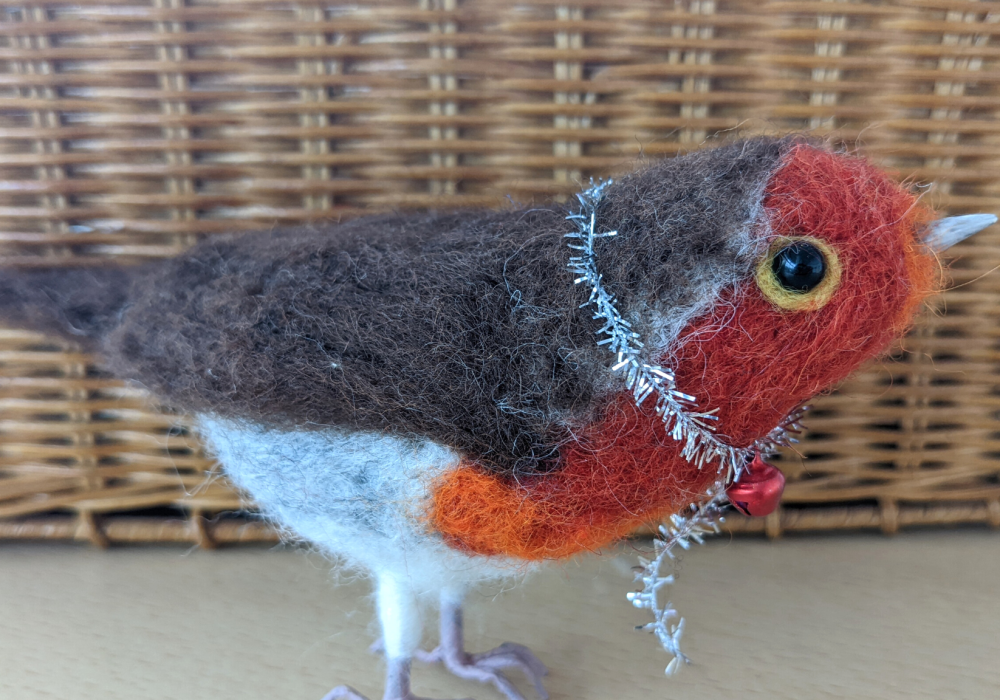 A needle felt sculpture of a robin, it has a string of silver tinsel around it's neck with a red bell.
