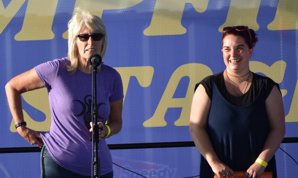 Ann and Sarah from the TWP on an outdoor stage. Ann is standing at a microphone.