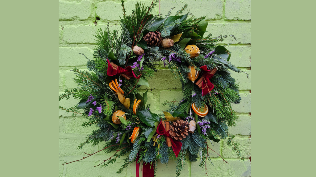 A photo of a lush green Christmas wreath, it is set against a pale green backdrop and has various decorations scattered throughout including pine cones, orange slices, and ribbon.