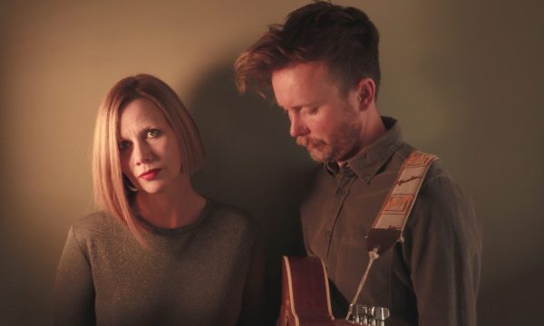 The band Megson stand in front of a dimly lit backdrop. On the left, a blonde female presenting person stares into the camera, and on the right a male presenting person holding a guitar looks down at the ground.