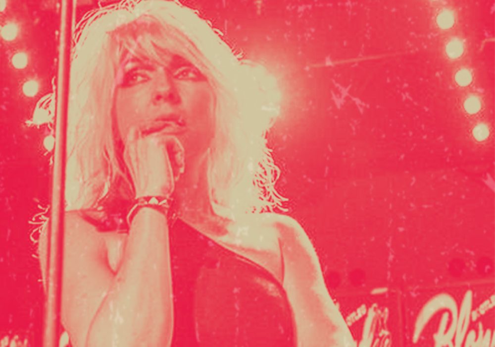 Bootleg Blondie frontwoman Debbie Harris stands in the centre of the image, only her head and shoulders are visible. She is looking off to the left of the camera, with her hand up against her mouth. The image has a red overlay.