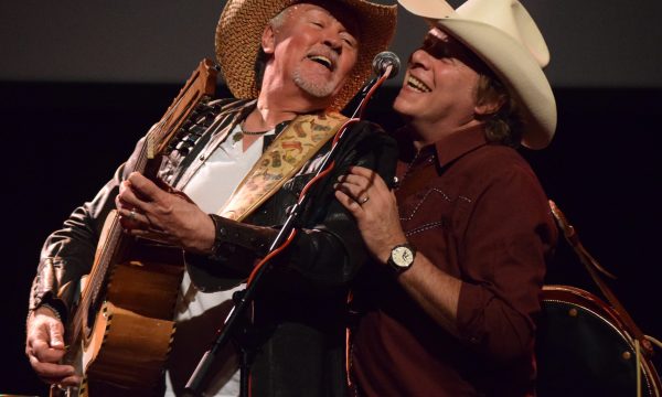 Two male musicians are both singing into a microphone, one man holds an acoustic guitar, both are wearing cowboy hats.