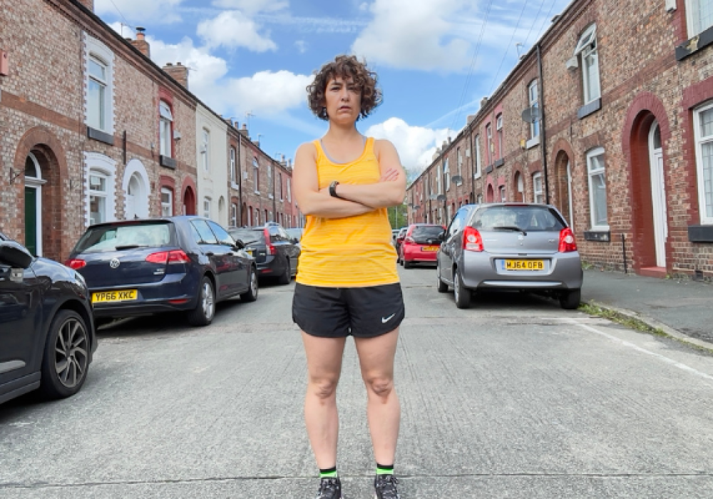 Jenni, wearing a yellow vest, black shorts, and black running shoes, stands with her arms folded in the middle of a road with terraced houses and cars on either side.