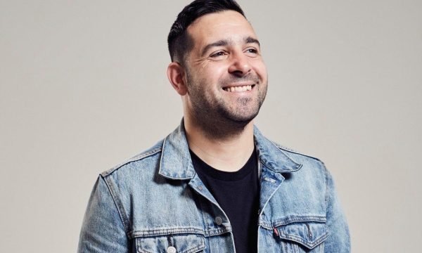 Comedian Danny McLoughlin, he is in front of a grey backdrop, and is grinning at something off to the right of the camera.