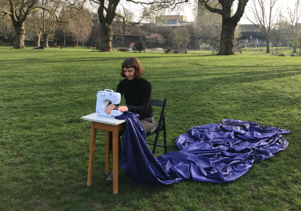 Natasha sits outside in an open, grassed area. She is sitting on a chair using a white sewing machine on a small table in front of her. A swathe of dark blue material fans out behind her from the sewing machine.