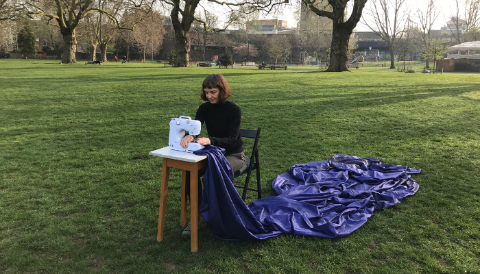 Natasha sits outside in an open, grassed area. She is sitting on a chair using a white sewing machine on a small table in front of her. A swathe of dark blue material fans out behind her from the sewing machine.