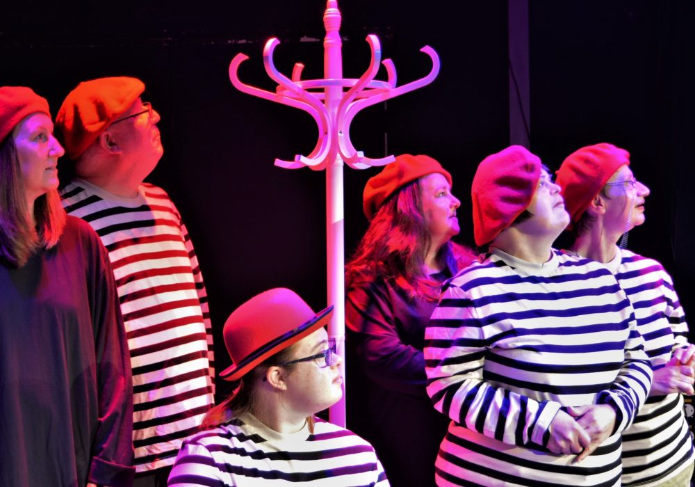 Six actors all looking up in the same direction, gathered around a tall white hat stand. Four are wearing stripy white and black tops, and two are wearing plain, dark coloured tops. Five are wearing matching red berets and another a red bowler hat.