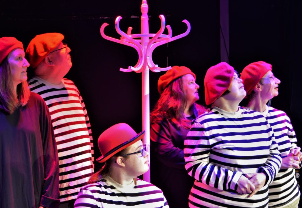 Six actors all looking up in the same direction, gathered around a tall white hat stand. Four are wearing stripy white and black tops, and two are wearing plain, dark coloured tops. Five are wearing matching red berets and another a red bowler hat.