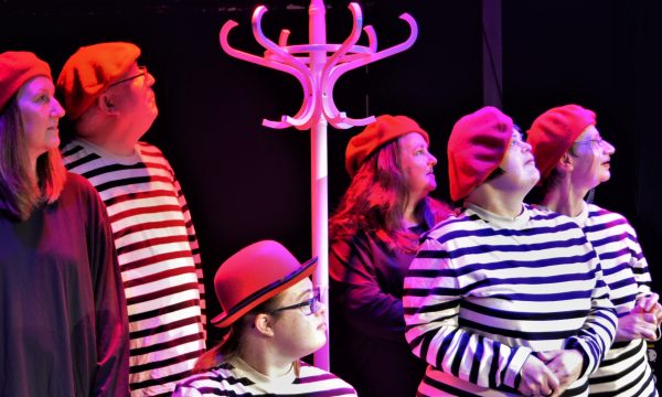 Six actors all looking up in the same direction, gathered around a tall white hat stand. Four are wearing stripy white and black tops, and two are wearing plain, dark coloured tops. Five are wearing matching red berets and another a red bowler hat.