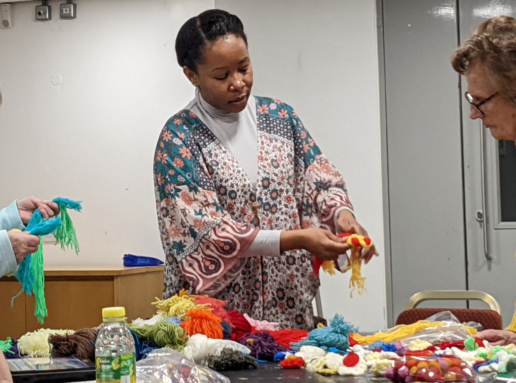 A photo of artist Jane, she is displaying some recycled wool jewelry to a class participant. The table in front of her is covered in multicoloured balls of wool. Just out of shot, another class participant is holding their own wool necklace made from blue and green wool.