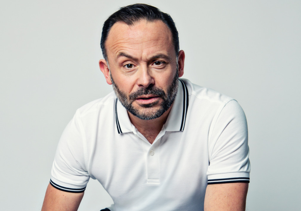 A headshot of comedian Geoff Norcott, he is wearing a white polo shirt and is looking intently into the camera with one eyebrow raised.