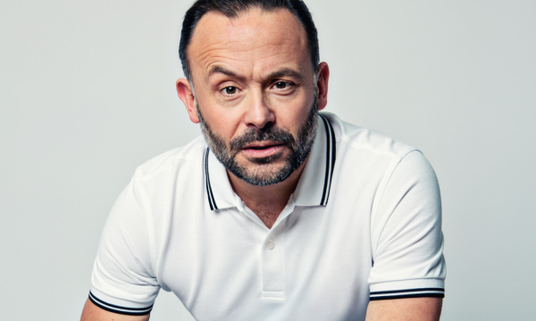 A headshot of comedian Geoff Norcott, he is wearing a white polo shirt and is looking intently into the camera with one eyebrow raised.