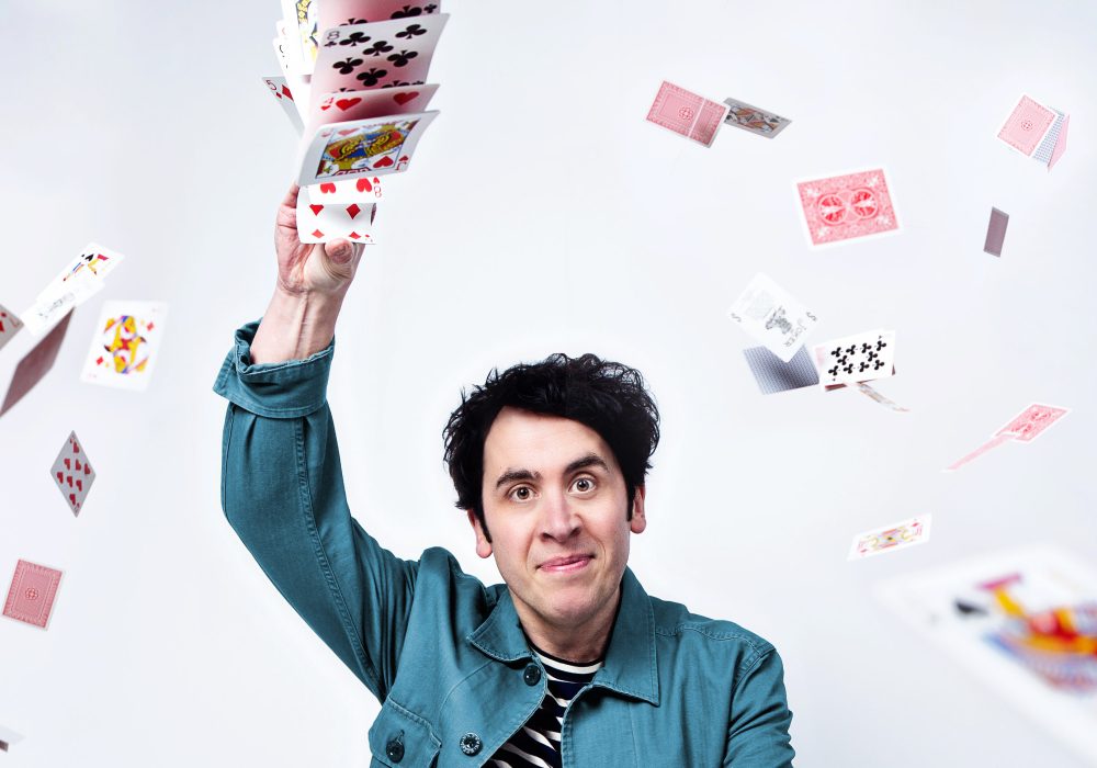 Comedian Pete Firman stands with one arm raised holding a stack of playing cards, more playing cards are flying around him