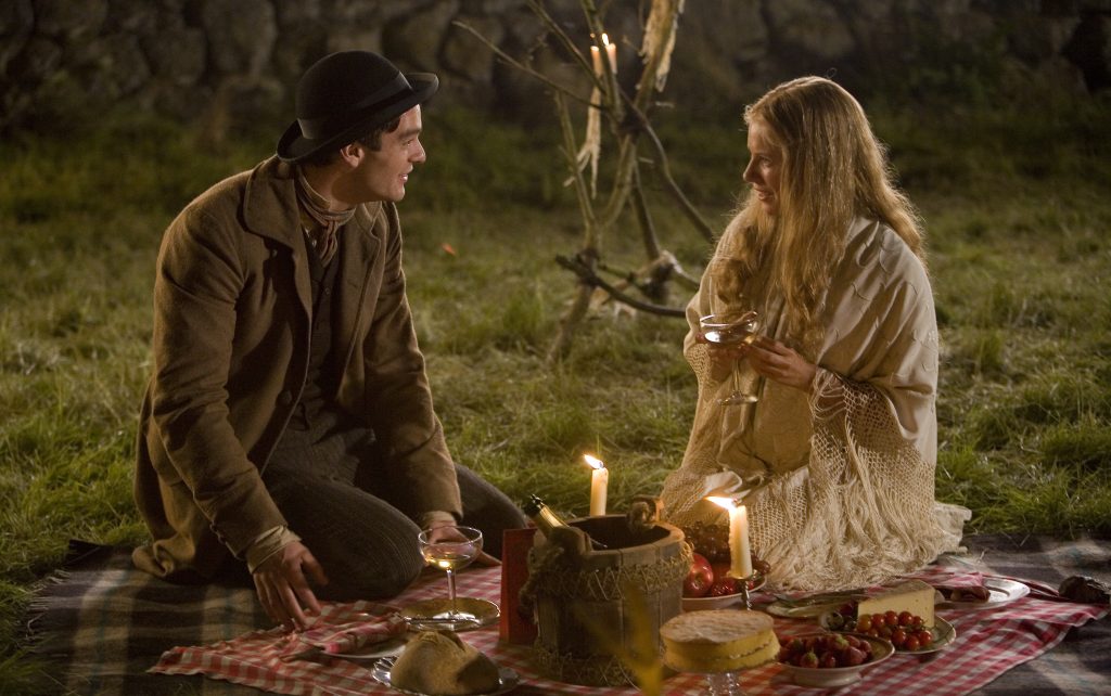 Image from the film Stardust that shows actor Charlie Cox as Tristan and Claire Danes as Yvaine. They are sat on a picnic blanket smiling at each other. They are surrounded by fruit, bread, and cake. There is a wooden bucket with a bottle of Champagne, and two lit candles on candlesticks.