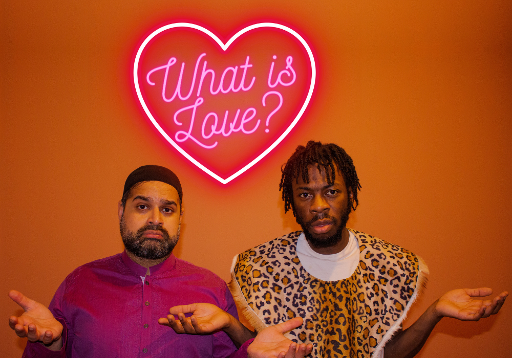 Umar Butt and Alberto Dumba stand side by side. They've both got their arms out, palms up, with questioning looks on their faces. Neon text reading 'What is Love' sits inside a neon heart shape.