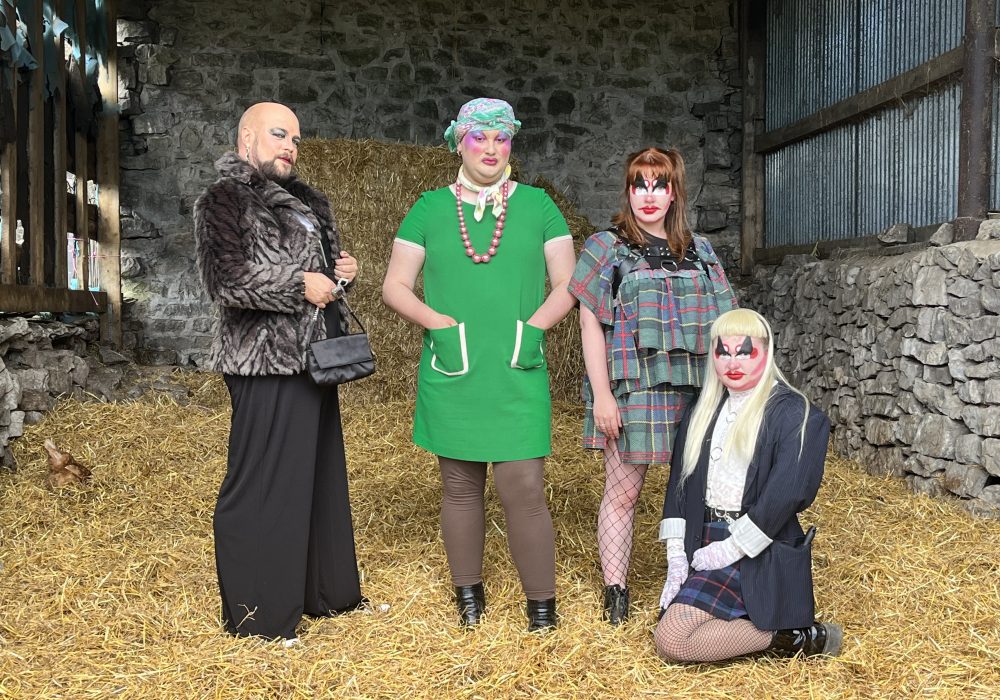 Four performers in drag, three standing and one kneeling on the floor of the straw filled barn they're in.