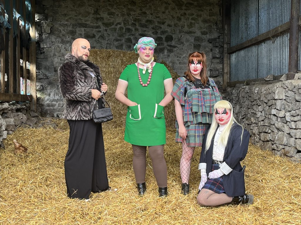 Four performers in drag, three standing and one kneeling on the floor of the straw filled barn they're in.