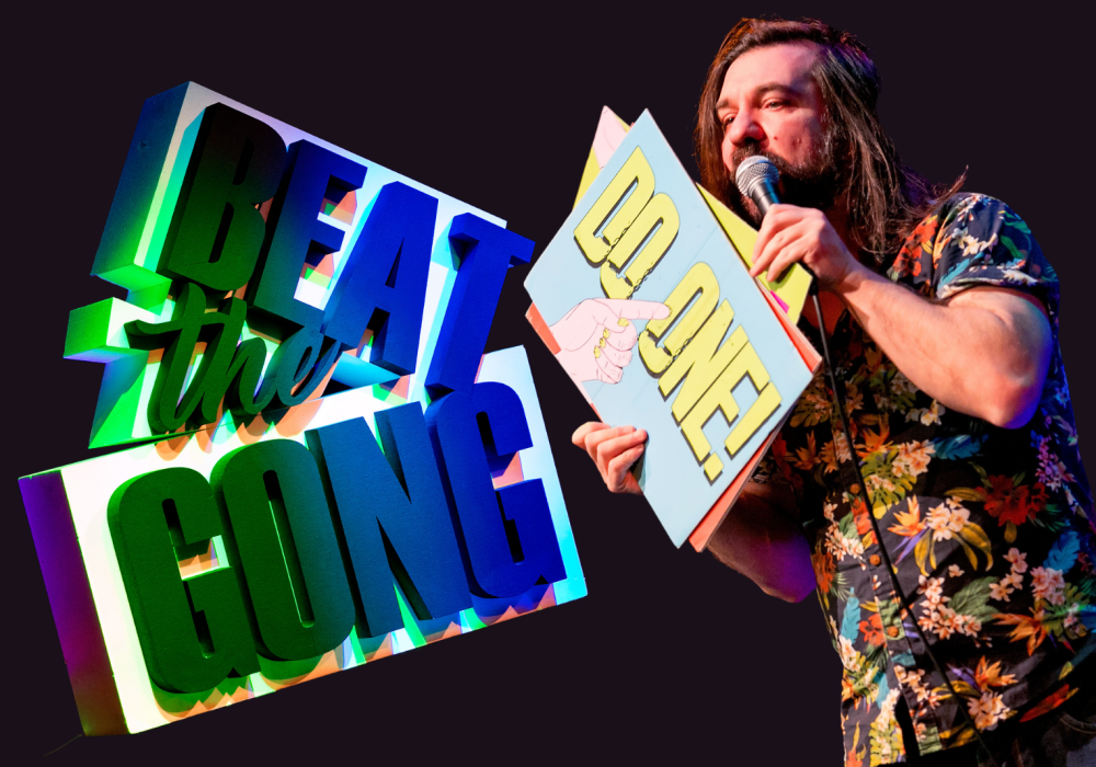 Comedian Matt Reed is speaking into a microphone and holding up a yellow and blue sign which reads "Do one." Next to him is the Beat the Gong logo.