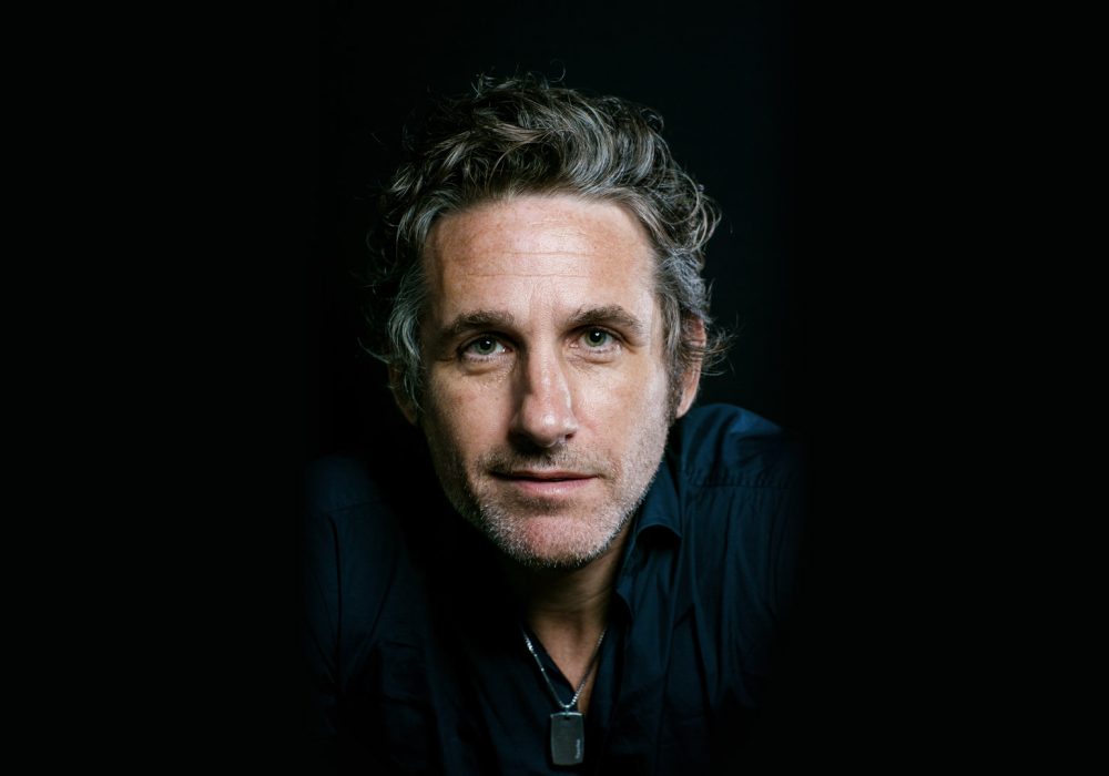 Comedian Tom Stade, he is leaning towards the camera and staring intently down the lens, the background is black.