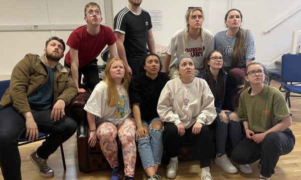 A cast of 10 students (most seated, some standing) in rehearsals for the show - they are all looking upwards in the same direction at something out of shot.