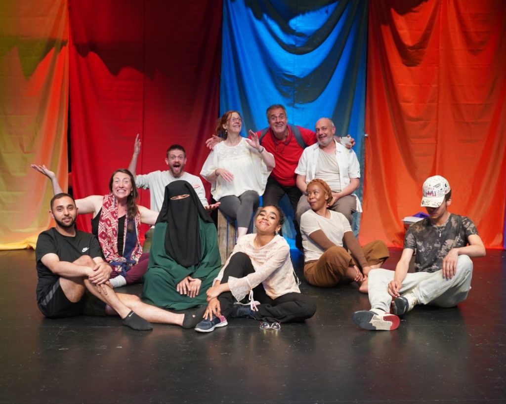 Alt: image of a group of people of various ethnicities sat on a theatre stage smiling at the camera, in the background there are number of colourful cloth banners hanging down.