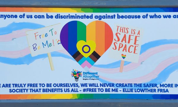 Free To Be Me mural, with trans colours and a placard illustration stating 'This is a safe space'. Text on the mural reads 'Any one of us can be discriminated against because of who we are. Until we are truly free to be ourselves, we will never create the safer, more inclusive society that benefits us all - #FreeToBeMe' - Ellie Lowther FRSA