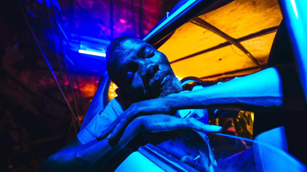 A man leans out of a car window, his arms rest on the doorframe and his eyes are closed. The lighting is blue.