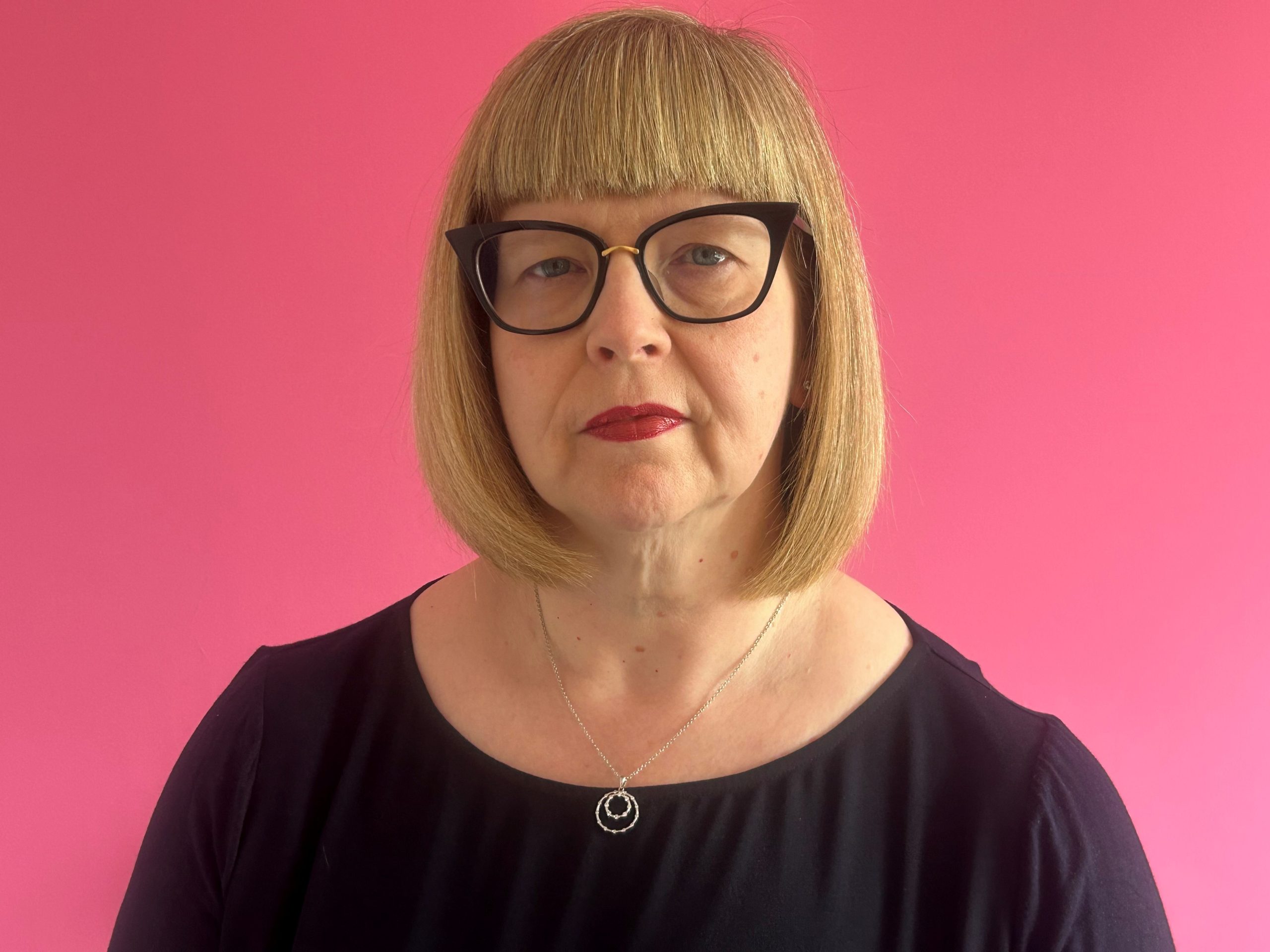 Photograph of Annabel Turpin, a woman in her early 50s, with a blond bob cut. She is wearing black glasses, red lipstick, and a dark purple top, and is standing in front of a pink background.